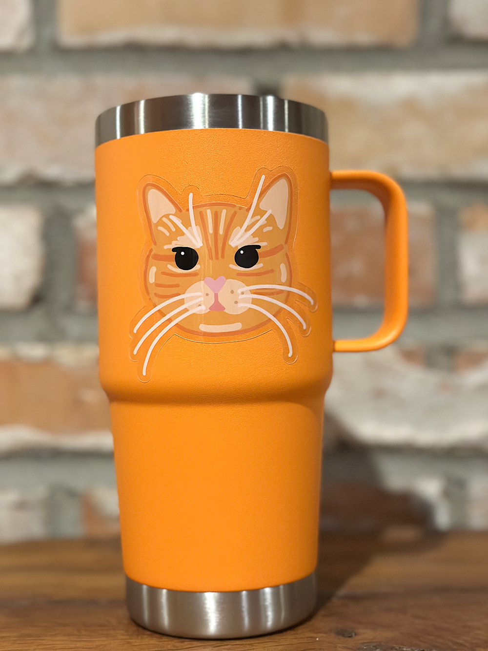 Orange travel mug with an orange cat face design on sticker and the sticker has no white border around the cat artwork. Transparent border sticker of cat. Background is a wooden surface