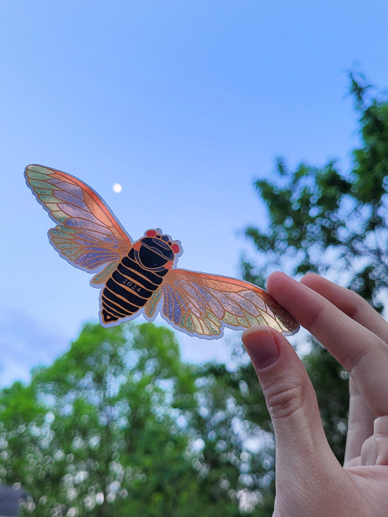 Hand holding a colorful cicada glitter sticker against a blue sky with trees in the background